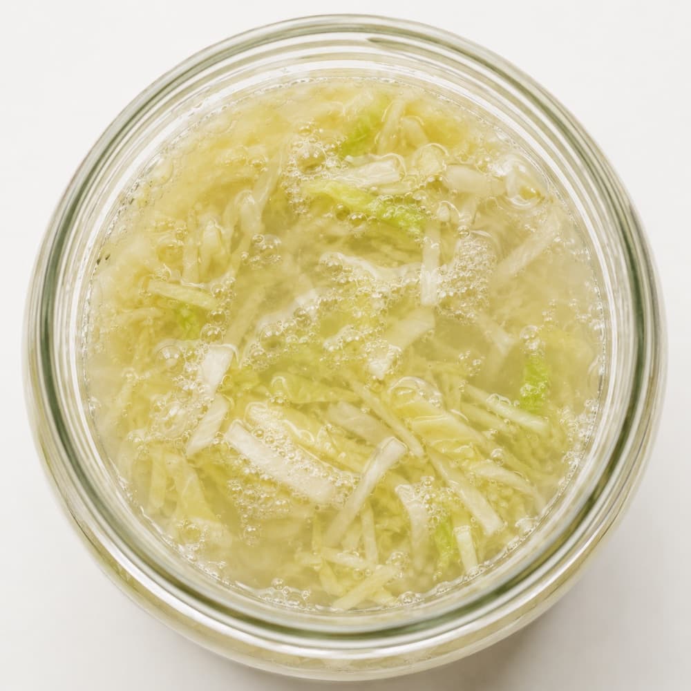 A healthy, active sauerkraut ferment: bubbles rising through a cloudy brine with the shredded cabbage fully submerged.